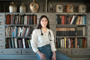 A woman sits in front of a bookshelf, surrounded by various vases and jugs, reflecting a serene indoor setting.